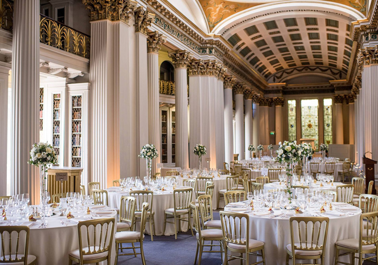 View of wedding meal tables with bouquets of white flowers in tapered vases and gold chairs