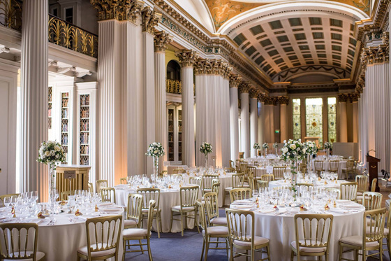 View of wedding meal tables with bouquets of white flowers in tapered vases and gold chairs